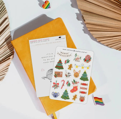 Two sheets of Christmas-themed stickers on a yellow notebook with a rainbow sticker and decorative paper in the background.